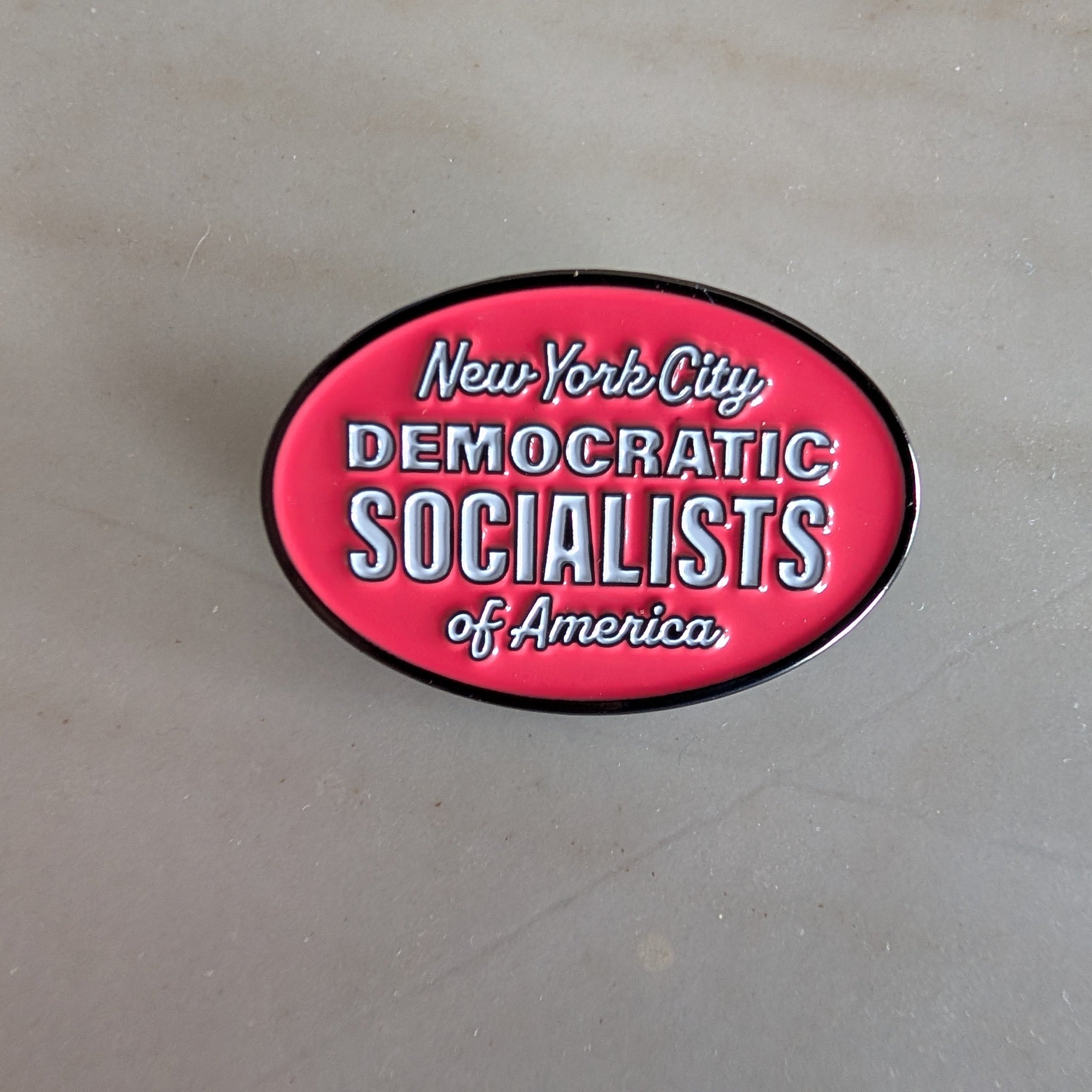 Pinkish red oval lapel pin with 'New York City Democratic Socialists of America' text on a gray background